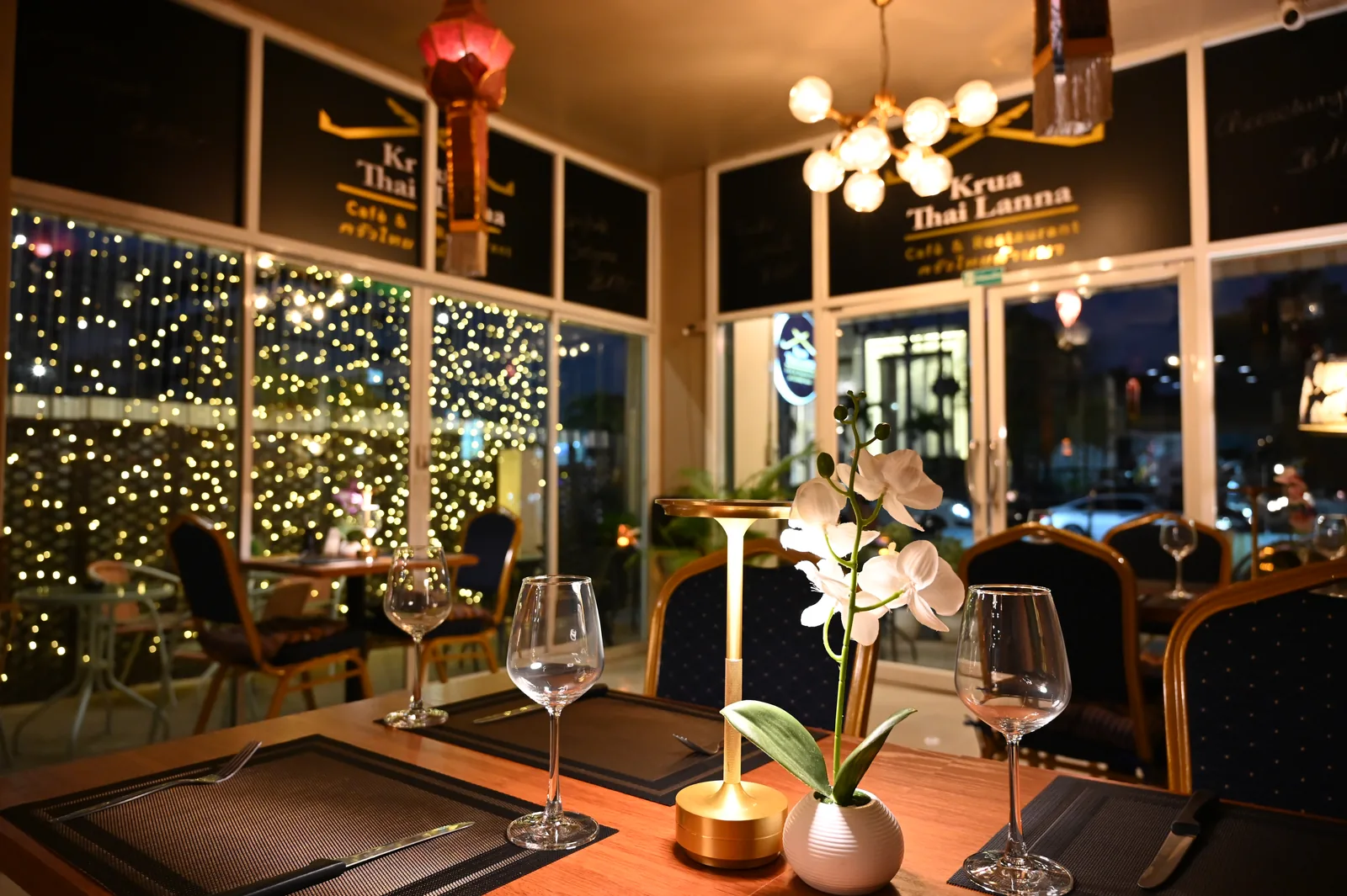 A candlelit table for two set with wine glasses and an orchid beside a window of twinkling lights at a restaurant in Thailand.